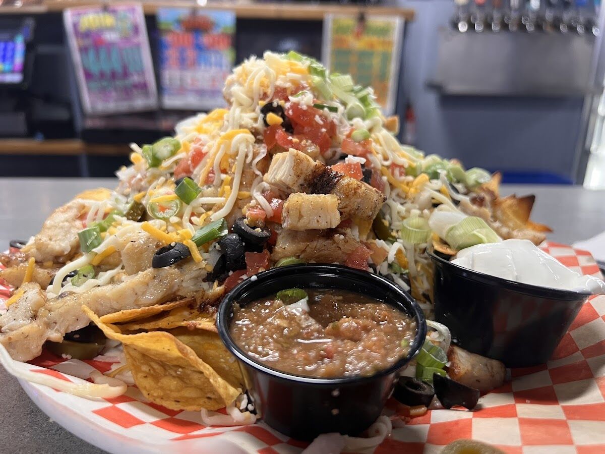 Nachos Supreme at Just Chill Pub & Grill — a mountain of tortilla chips with grilled chicken, melted cheese, pico de gallo, black olives, jalapeños and green onion, with salsa and sour cream, the beer taps behind.